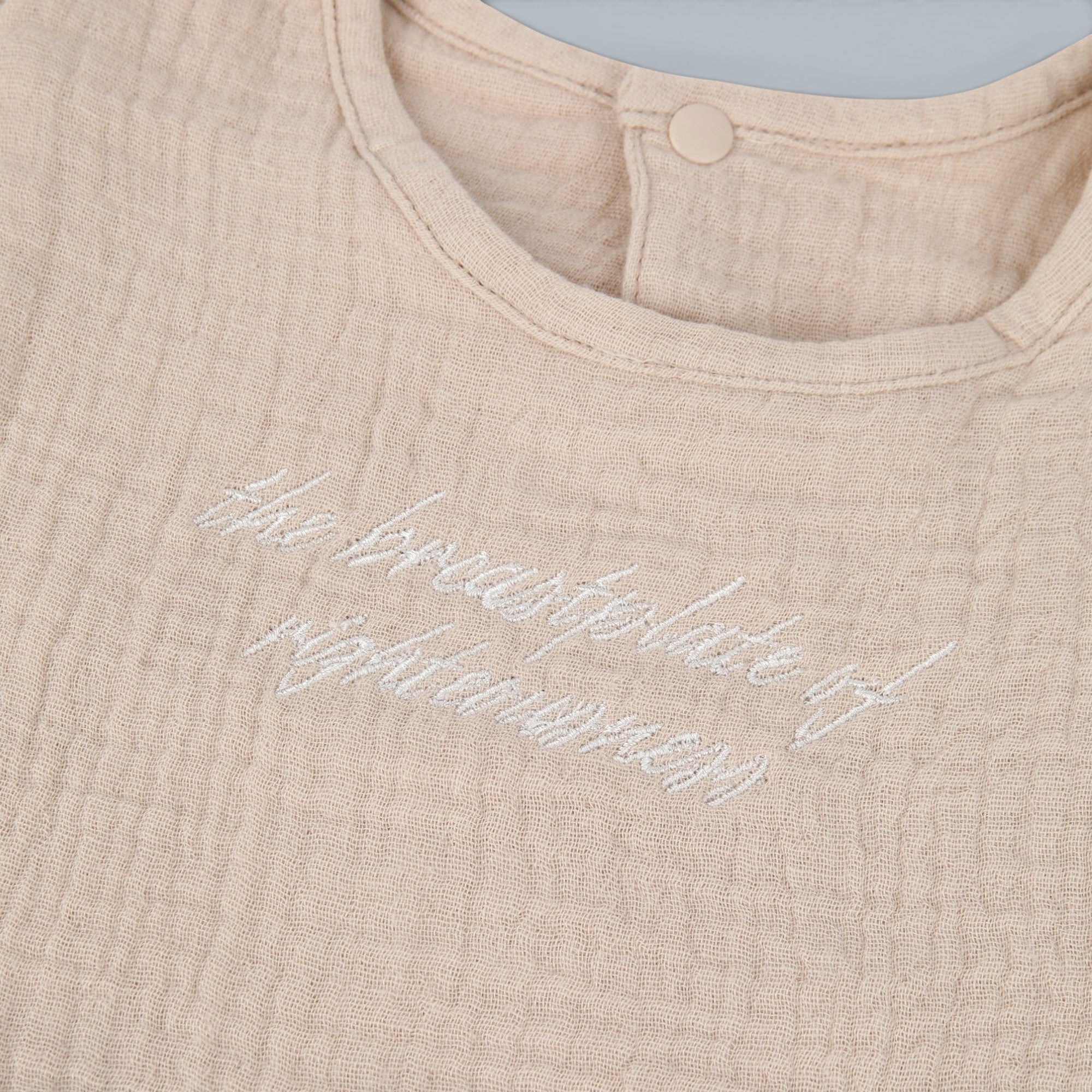 Shalom Living beige cotton gauze ruffle bib with embroidered text "the breastplate of righteousness" and snap closure.