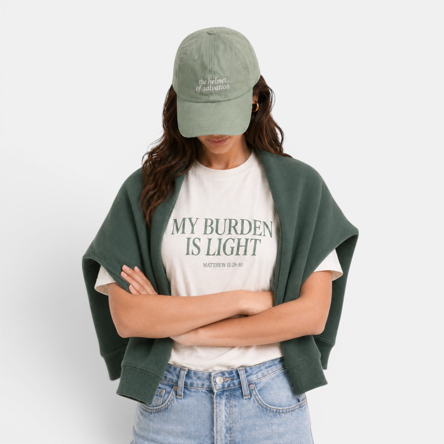 Shalom Living cream relaxed fit t-shirt with green text "MY BURDEN IS LIGHT MATTHEW 11:28-30". Model wears a green cap and green sweater draped over shoulders.