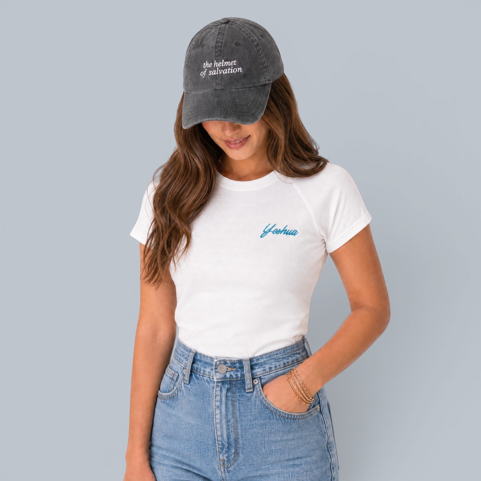 Shalom Living white fitted t-shirt with blue "Yeshua" embroidery on the chest, worn by a woman with jeans and a grey baseball cap.