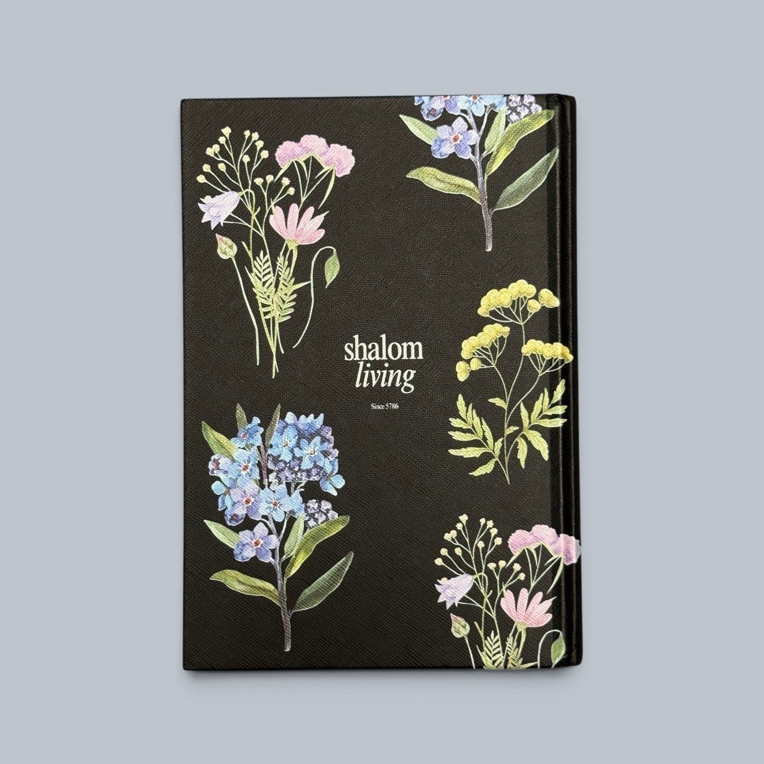 Shalom Living black floral journal book with lined paper pages and softcover finish. Features delicate watercolor illustrations of forget-me-nots, pink wildflowers, and yellow blossoms.